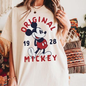 May include: Ivory-colored t-shirt featuring a classic Mickey Mouse graphic. The design includes the words "ORIGINAL MICKEY 1928" in red and navy blue. The shirt is a relaxed fit, with the word "IVORY" printed at the bottom.