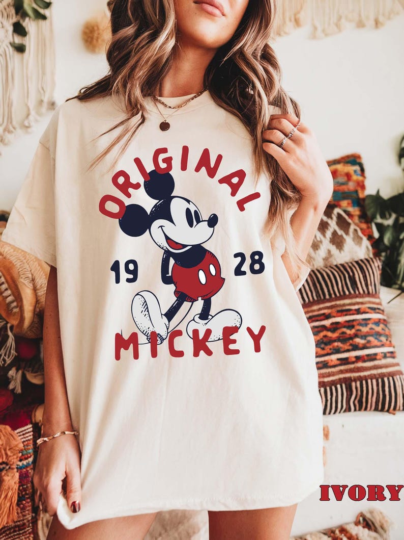 May include: Ivory-colored t-shirt featuring a classic Mickey Mouse graphic. The design includes the words "ORIGINAL MICKEY 1928" in red and navy blue. The shirt is a relaxed fit, with the word "IVORY" printed at the bottom.