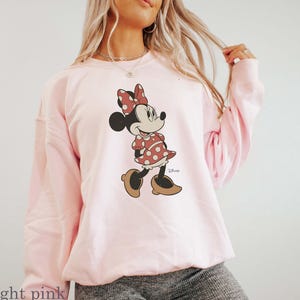 May include: Light pink sweatshirt featuring a Minnie Mouse graphic. The cartoon character is depicted in a red polka dot dress, matching bow, and brown shoes. The Disney logo is visible on the bottom right of the graphic. The sweatshirt is a soft, casual style.
