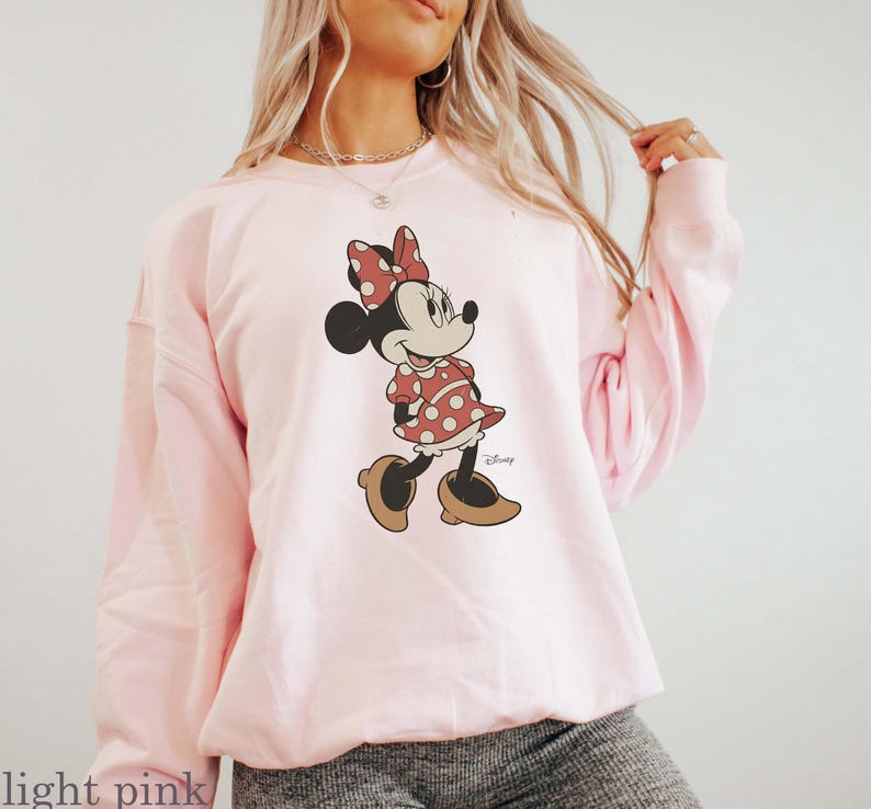 May include: Light pink sweatshirt featuring a Minnie Mouse graphic. The cartoon character is depicted in a red polka dot dress, matching bow, and brown shoes. The Disney logo is visible on the bottom right of the graphic. The sweatshirt is a soft, casual style.
