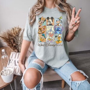 Op de afbeelding: Saliegroen T-shirt met een rasterontwerp met Disney-personages zoals Mickey Mouse, Minnie Mouse, Donald Duck en Stitch. De woorden "Walt Disney World" staan eronder gedrukt. Het shirt wordt gedragen met lichtblauwe ripped jeans.