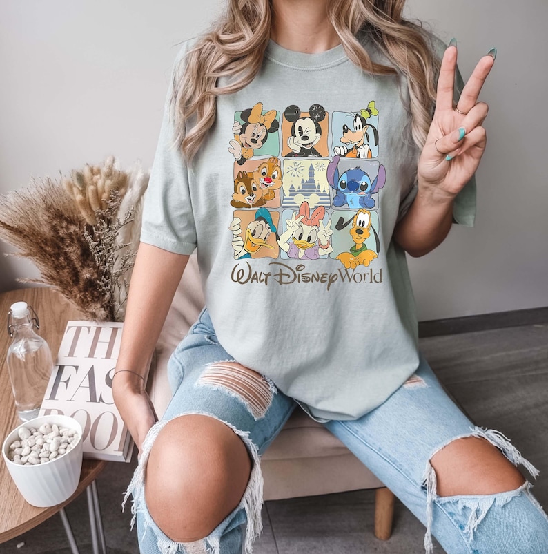 Op de afbeelding: Saliegroen T-shirt met een rasterontwerp met Disney-personages zoals Mickey Mouse, Minnie Mouse, Donald Duck en Stitch. De woorden "Walt Disney World" staan eronder gedrukt. Het shirt wordt gedragen met lichtblauwe ripped jeans.