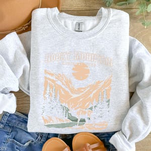 Pu&ograve; includere: Una felpa grigia con una stampa grafica di una catena montuosa con il testo "Rocky Mountain" in color pesca. La felpa &egrave; realizzata in un materiale morbido e confortevole.