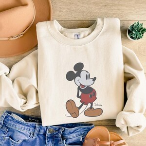 May include: Cream-colored sweatshirt featuring a graphic of Mickey Mouse in his classic pose. The cartoon character is depicted in black, red, and brown. The sweatshirt is paired with blue denim shorts, a tan hat, and sandals.