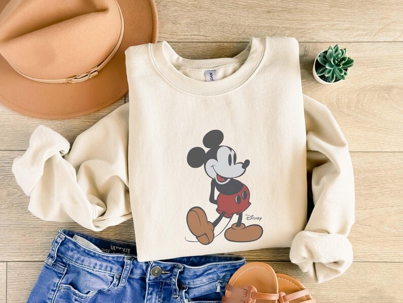 May include: Cream-colored sweatshirt featuring a graphic of Mickey Mouse in his classic pose. The cartoon character is depicted in black, red, and brown. The sweatshirt is paired with blue denim shorts, a tan hat, and sandals.