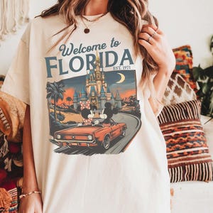 May include: Ivory-colored t-shirt featuring a graphic of Mickey and Minnie Mouse in a red convertible driving towards a castle. The shirt has the text "Welcome to Florida EST. 1971" in blue and white.