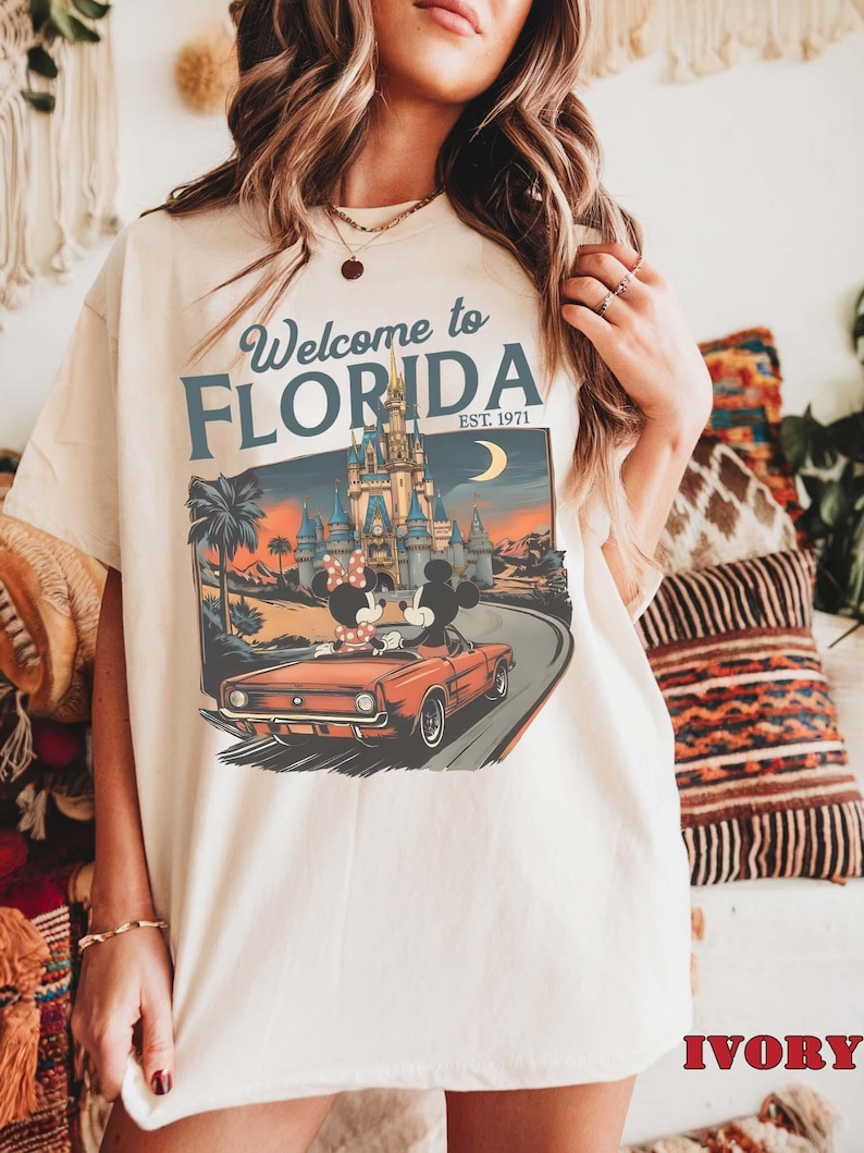May include: Ivory-colored t-shirt featuring a graphic of Mickey and Minnie Mouse in a red convertible driving towards a castle. The shirt has the text "Welcome to Florida EST. 1971" in blue and white.