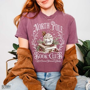 K&ouml;nnte beinhalten: Beerenfarbenes T-Shirt mit einem "North Pole Book Club"-Motiv. Das Design zeigt eine Tasse hei&szlig;e Schokolade, einen Lebkuchenmann und den Text "Hot Cocoa Served Daily". Das Shirt hat einen Rundhalsausschnitt.