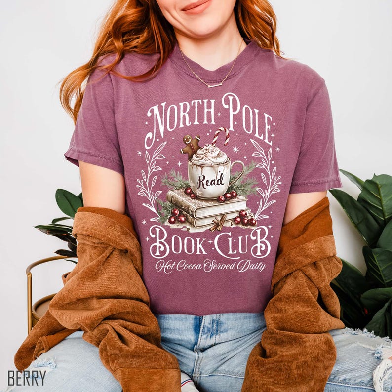 K&ouml;nnte beinhalten: Beerenfarbenes T-Shirt mit einem "North Pole Book Club"-Motiv. Das Design zeigt eine Tasse hei&szlig;e Schokolade, einen Lebkuchenmann und den Text "Hot Cocoa Served Daily". Das Shirt hat einen Rundhalsausschnitt.
