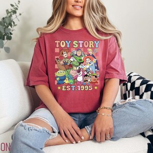 Może przedstawiać: Czerwony T-shirt z grafiką w stylu vintage postaci z Toy Story, w tym Woody, Buzz Astral i Rex. Grafika jest otoczona kratką i tekstem "Toy Story Est. 1995".