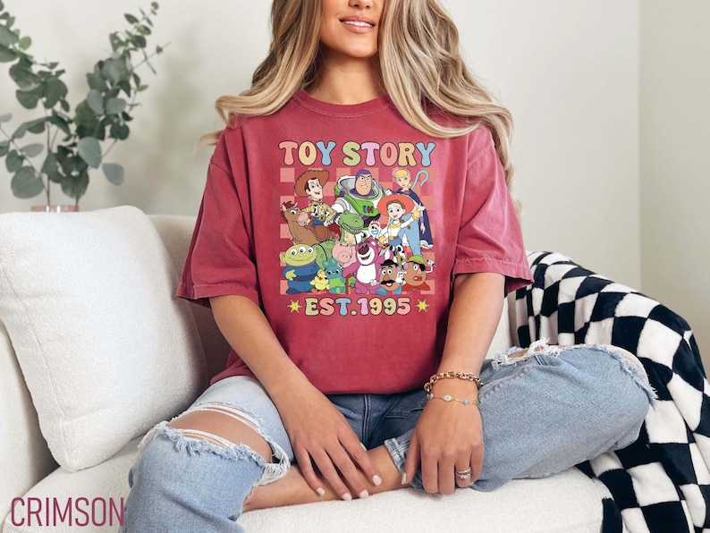 Może przedstawiać: Czerwony T-shirt z grafiką w stylu vintage postaci z Toy Story, w tym Woody, Buzz Astral i Rex. Grafika jest otoczona kratką i tekstem "Toy Story Est. 1995".