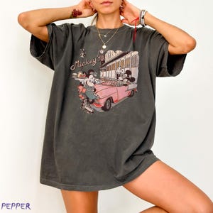 May include: Dark grey t-shirt featuring a vintage-style graphic of Mickey and Minnie Mouse in a pink convertible car in front of a diner. The shirt has the text "Mickey's Cafe 50's" in a script font. The shirt is oversized.