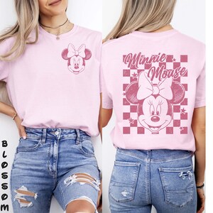 May include: Light pink t-shirt featuring a Minnie Mouse graphic on the front and back. The front graphic is a small Minnie Mouse head with a bow. The back graphic features a larger Minnie Mouse head with a bow, the words "Minnie Mouse", and a checkered background.