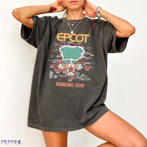 May include: Dark gray t-shirt with the word "EPCOT" in orange and a map design. Below the map, the text reads "DRINKING MAP". The shirt has short sleeves and a relaxed fit.
