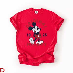 May include: A red t-shirt featuring a graphic of Mickey Mouse with the words "ORIGINAL 1928 MICKEY" in a circular design. The cartoon character is depicted in black, white, and red. The shirt is a solid red color.