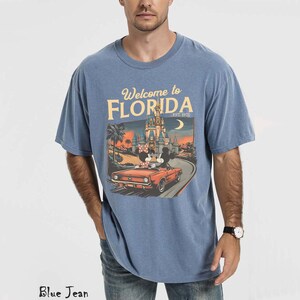 May include: Blue short-sleeved t-shirt with the text "Welcome to Florida" and an illustration of a car with Mickey and Minnie Mouse in front of a castle. The shirt is a light blue color.