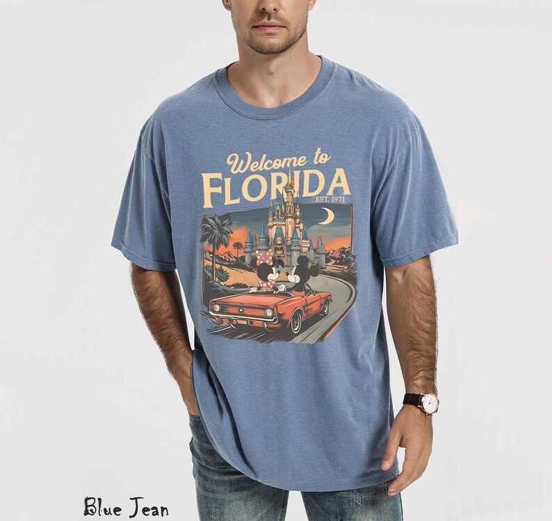 May include: Blue short-sleeved t-shirt with the text "Welcome to Florida" and an illustration of a car with Mickey and Minnie Mouse in front of a castle. The shirt is a light blue color.