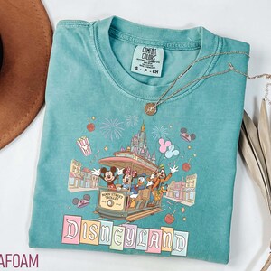 May include: Seafoam green t-shirt featuring a Disneyland Main Street Trolley design with Mickey Mouse, Minnie Mouse, Donald Duck, and Goofy. The shirt has a crew neck and short sleeves. A gold necklace with a pendant is also visible.