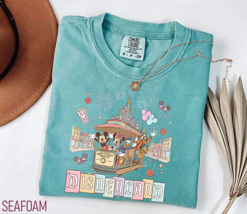 May include: Seafoam green t-shirt featuring a Disneyland Main Street Trolley design with Mickey Mouse, Minnie Mouse, Donald Duck, and Goofy. The shirt has a crew neck and short sleeves. A gold necklace with a pendant is also visible.