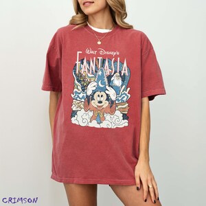 May include: Crimson red t-shirt with a graphic of Mickey Mouse in a wizard hat from Walt Disney's Fantasia. The design includes the film's title and imagery of clouds and stars. The shirt has short sleeves and a relaxed fit.