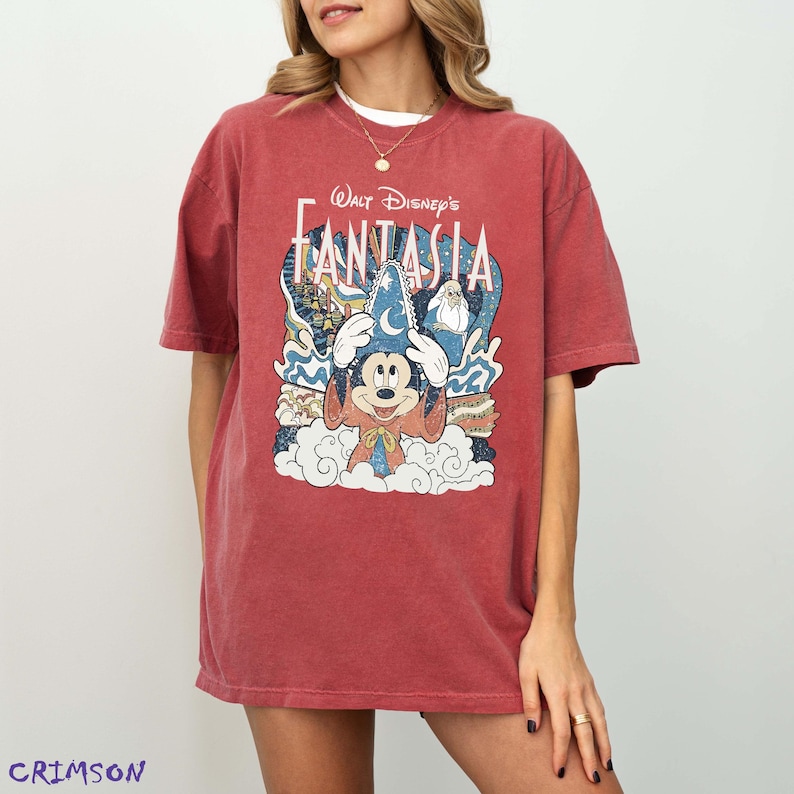 May include: Crimson red t-shirt with a graphic of Mickey Mouse in a wizard hat from Walt Disney's Fantasia. The design includes the film's title and imagery of clouds and stars. The shirt has short sleeves and a relaxed fit.
