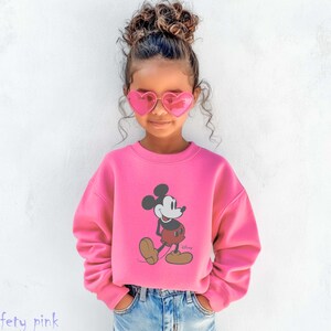 May include: A child wearing a safety pink sweatshirt featuring a Mickey Mouse graphic. The sweatshirt is paired with blue jeans and pink heart-shaped sunglasses. The word "Safety pink" is printed at the bottom of the image.