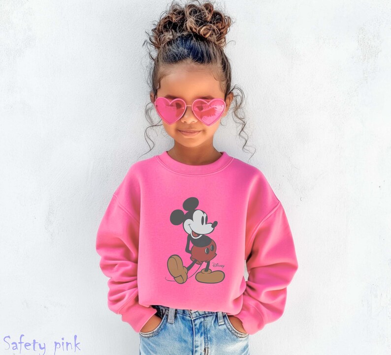 May include: A child wearing a safety pink sweatshirt featuring a Mickey Mouse graphic. The sweatshirt is paired with blue jeans and pink heart-shaped sunglasses. The word "Safety pink" is printed at the bottom of the image.