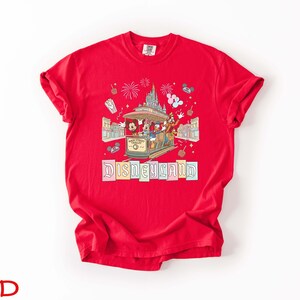 May include: A red t-shirt featuring a Disneyland design. The graphic includes Mickey Mouse, Donald Duck, and Goofy riding a trolley, with fireworks and the Disneyland logo. The shirt is made of a soft material.