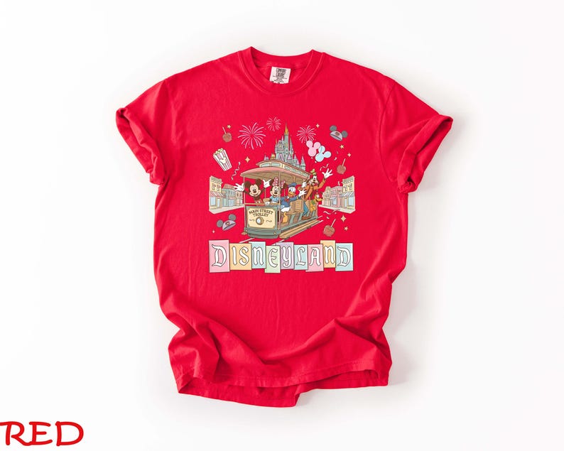 May include: A red t-shirt featuring a Disneyland design. The graphic includes Mickey Mouse, Donald Duck, and Goofy riding a trolley, with fireworks and the Disneyland logo. The shirt is made of a soft material.