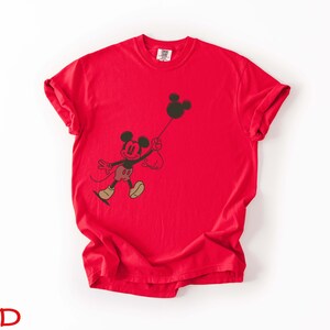 May include: A vibrant red t-shirt with a black graphic of Mickey Mouse holding a balloon. The cartoon character is wearing red shorts, yellow shoes, and a black face. The word "Smile" is written on the shirt.