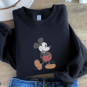 May include: Black crewneck sweatshirt featuring a Mickey Mouse graphic. The cartoon character is depicted in a classic pose, wearing red shorts and yellow shoes. The word "Disney" is printed below the character. The word "BLACK" is printed at the bottom.
