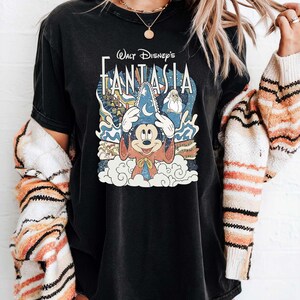 May include: Black t-shirt with the text "Walt Disney's Fantasia" and a graphic of Mickey Mouse as a sorcerer. The design includes clouds, stars, and characters from the film. The shirt is worn by a person.