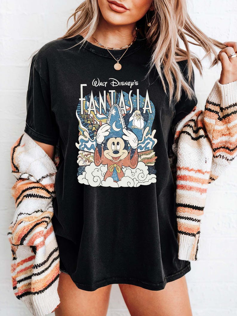 May include: Black t-shirt with the text "Walt Disney's Fantasia" and a graphic of Mickey Mouse as a sorcerer. The design includes clouds, stars, and characters from the film. The shirt is worn by a person.