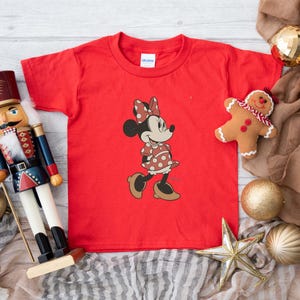 May include: A red t-shirt featuring a Minnie Mouse graphic. The cartoon character is wearing a polka dot dress and bow. The image also includes a toy nutcracker, a gingerbread man, and Christmas ornaments.