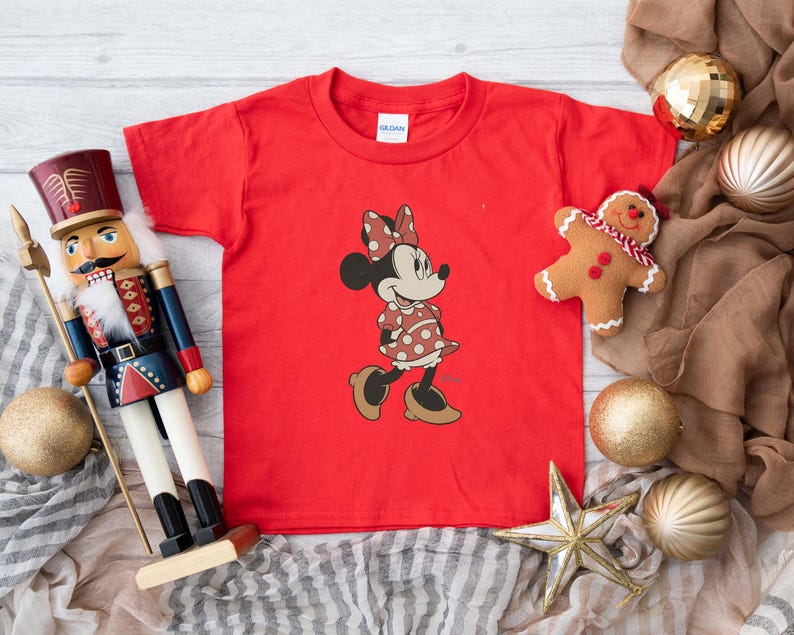 May include: A red t-shirt featuring a Minnie Mouse graphic. The cartoon character is wearing a polka dot dress and bow. The image also includes a toy nutcracker, a gingerbread man, and Christmas ornaments.