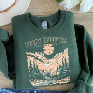 Pu&ograve; includere: Una felpa verde scuro con una stampa grafica di una catena montuosa e il testo "Rocky Mountain - National Park -" in bianco.