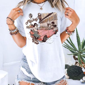 May include: White t-shirt featuring a vintage-style graphic of Mickey and Minnie Mouse. Mickey is driving a pink convertible, while Minnie is roller skating. The graphic includes the text "Mickey" and "Cafe 50's".