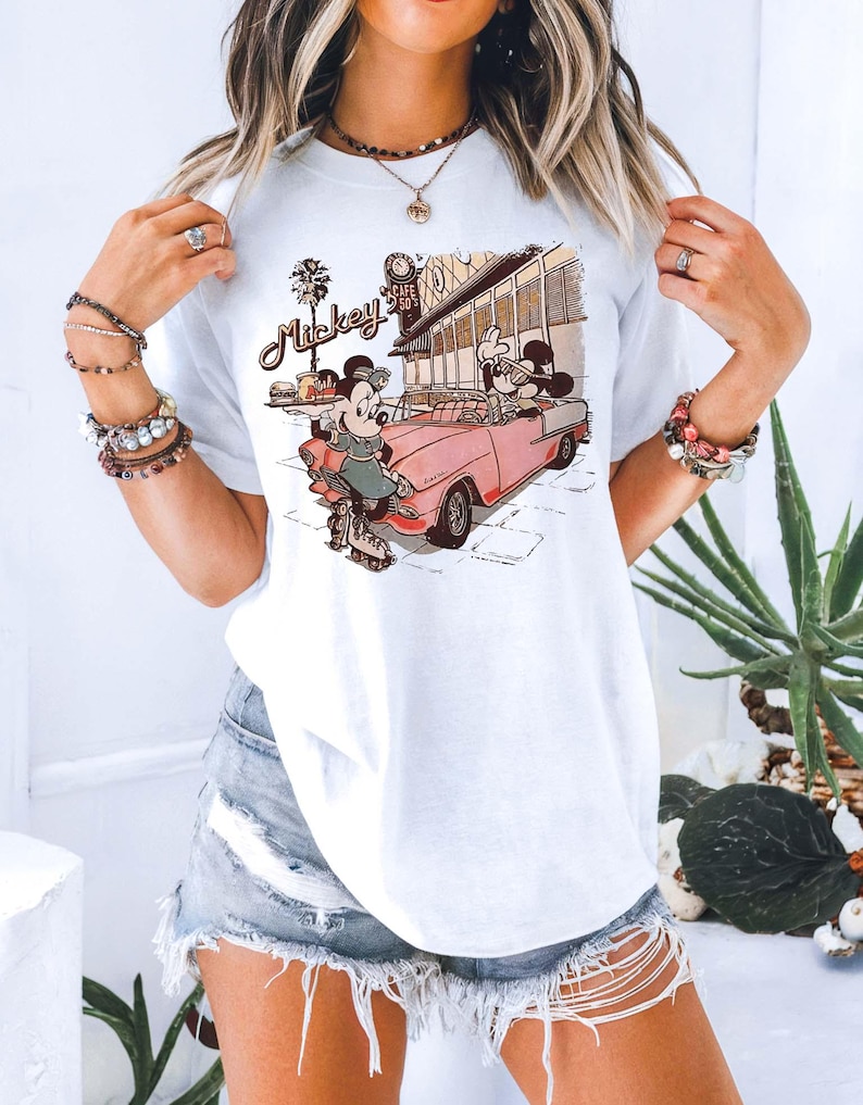 May include: White t-shirt featuring a vintage-style graphic of Mickey and Minnie Mouse. Mickey is driving a pink convertible, while Minnie is roller skating. The graphic includes the text "Mickey" and "Cafe 50's".
