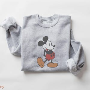 May include: A light gray sweatshirt featuring a graphic of Mickey Mouse in his classic pose. Mickey is depicted with black ears, a white face, red shorts, and brown shoes. The word "Disney" is printed on the bottom right.