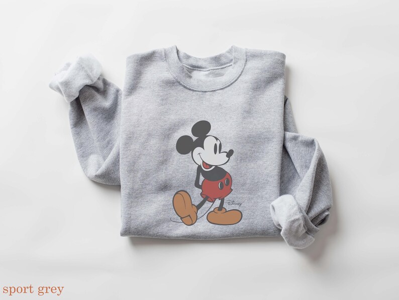 May include: A light gray sweatshirt featuring a graphic of Mickey Mouse in his classic pose. Mickey is depicted with black ears, a white face, red shorts, and brown shoes. The word "Disney" is printed on the bottom right.
