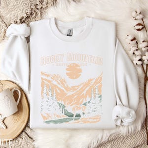 Pu&ograve; includere: Felpa bianca con una stampa grafica di una catena montuosa e il testo "Rocky Mountain National Park".