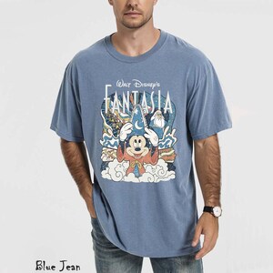 May include: A blue t-shirt with a graphic of Mickey Mouse in a sorcerer's hat from Walt Disney's Fantasia. The design includes clouds, stars, and other characters. The shirt is a solid blue colour.