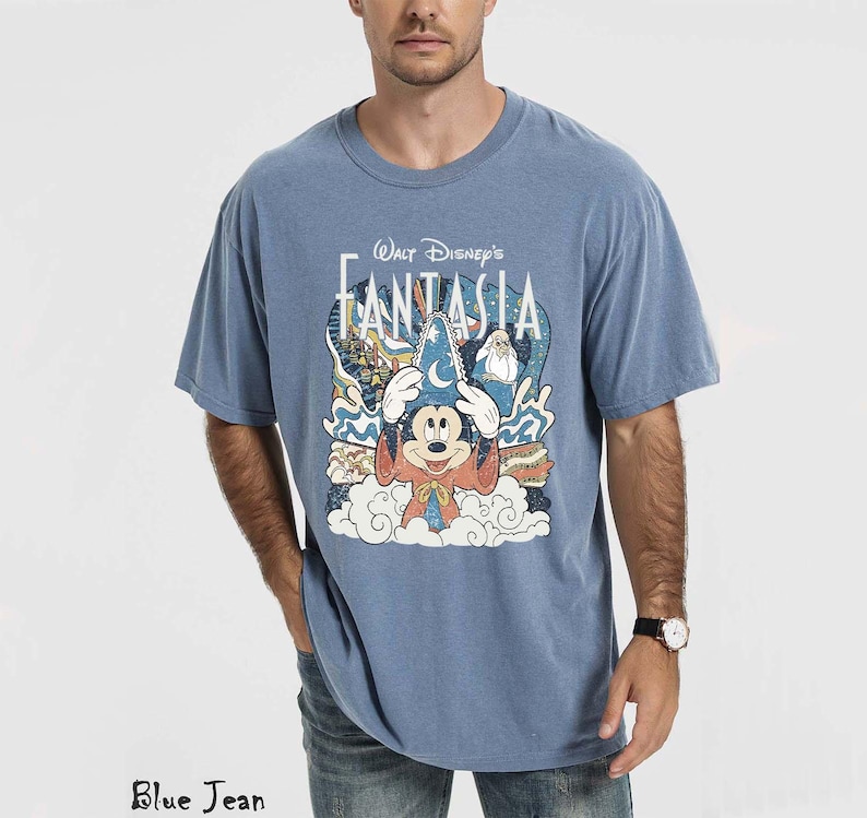 May include: A blue t-shirt with a graphic of Mickey Mouse in a sorcerer's hat from Walt Disney's Fantasia. The design includes clouds, stars, and other characters. The shirt is a solid blue colour.