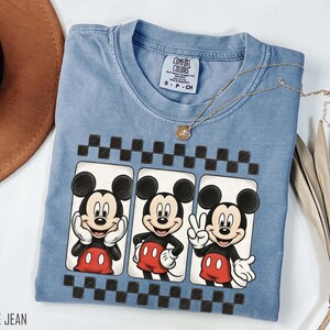 May include: A blue jean colored t-shirt featuring three Mickey Mouse illustrations in white rectangular frames with a black and white checkered border. The shirt has a crew neck and short sleeves. A gold necklace is draped across the shirt.