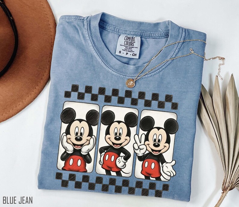May include: A blue jean colored t-shirt featuring three Mickey Mouse illustrations in white rectangular frames with a black and white checkered border. The shirt has a crew neck and short sleeves. A gold necklace is draped across the shirt.