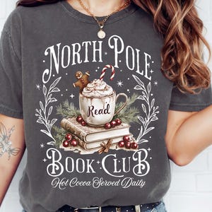 K&ouml;nnte beinhalten: Dunkelgraues T-Shirt mit festlichem Design. Die Grafik zeigt eine Tasse hei&szlig;e Schokolade mit Schlagsahne, einen Lebkuchenmann und eine Zuckerstange. Der Text lautet "North Pole Book Club" und "Hot Cocoa Served Daily".