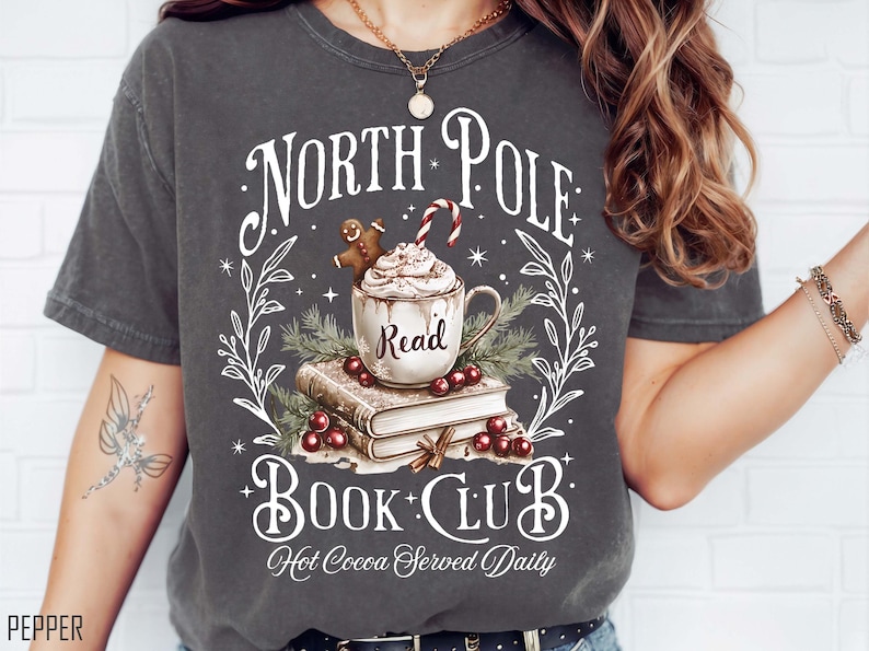 K&ouml;nnte beinhalten: Dunkelgraues T-Shirt mit festlichem Design. Die Grafik zeigt eine Tasse hei&szlig;e Schokolade mit Schlagsahne, einen Lebkuchenmann und eine Zuckerstange. Der Text lautet "North Pole Book Club" und "Hot Cocoa Served Daily".