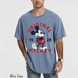 May include: A light blue t-shirt featuring a graphic of Mickey Mouse with the text "ORIGINAL 1928 MICKEY" in red. The shirt has a crew neck and short sleeves. The model is wearing blue jeans.