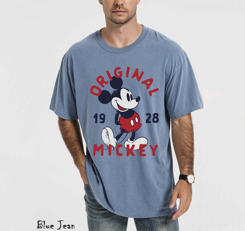 May include: A light blue t-shirt featuring a graphic of Mickey Mouse with the text "ORIGINAL 1928 MICKEY" in red. The shirt has a crew neck and short sleeves. The model is wearing blue jeans.