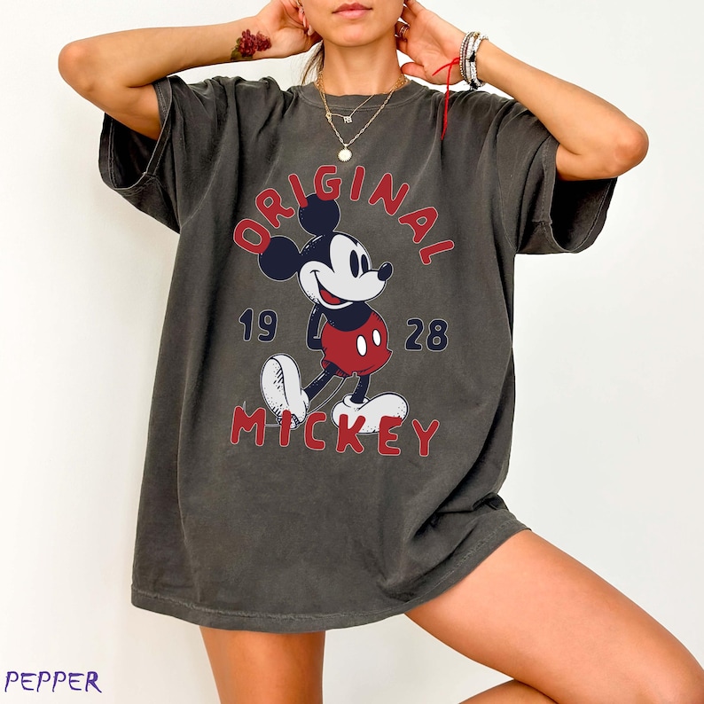 May include: Dark gray t-shirt featuring a classic Mickey Mouse graphic. The design includes the words "ORIGINAL MICKEY" in red, with the year "1928" below the word "ORIGINAL". The iconic cartoon character is depicted in his signature colors.